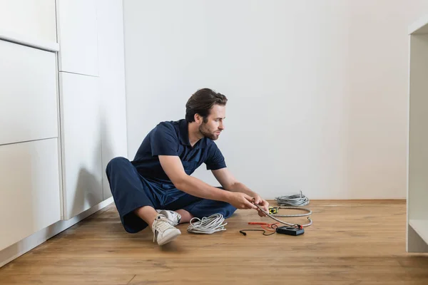 Electricista en uniforme sentado en el suelo en la cocina cerca de cables y probador eléctrico - foto de stock