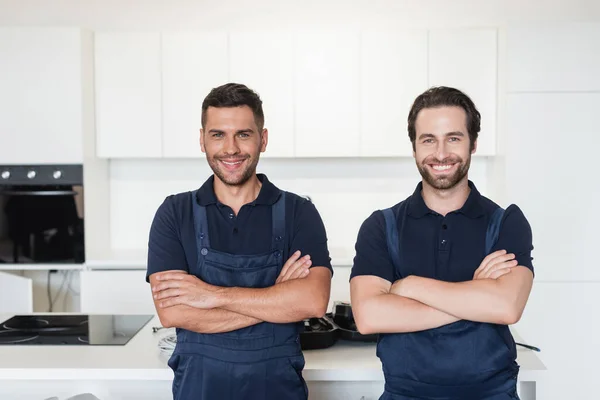 Happy handymen in uniform standing with crossed arms in kitchen — Stock Photo