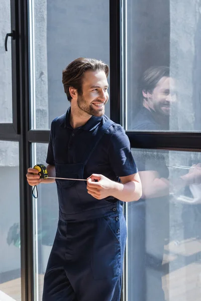 Happy handyman with measuring tape looking away near large windows — Stock Photo