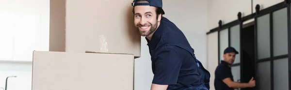 Happy mover smiling near cardboard packages and blurred colleague, banner — Stock Photo