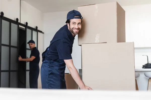 Happy mover smiling near carton boxes and colleague on blurred background — Stock Photo