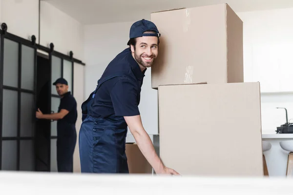 Feliz mover sorrindo perto de caixas de papelão e colega no fundo borrado — Fotografia de Stock