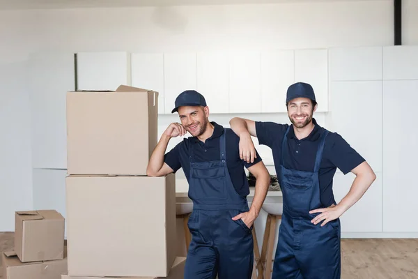 Gioiosi traslocatori sorridenti alla macchina fotografica vicino a confezioni di cartone in cucina — Foto stock