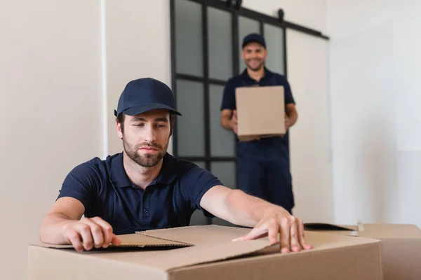 Arbeiter in Mütze packt Karton neben verschwommenem Kollegen aus — Stockfoto