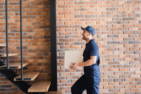 Side view of happy mover in uniform walking upstairs with cardboard box — Stock Photo