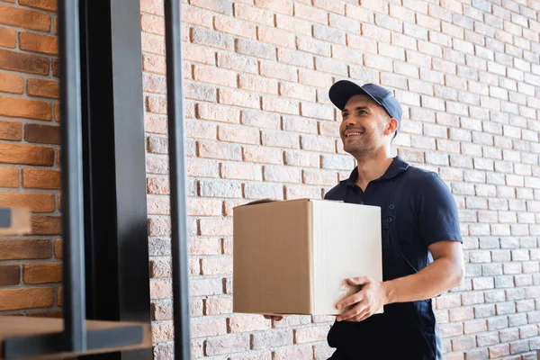 Cheerful delivery man holding cardboard package near brick wall — Stock Photo