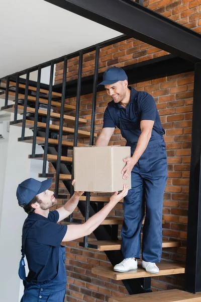 Workman giving carton box to smiling mover standing on stairs in apartment — Stock Photo