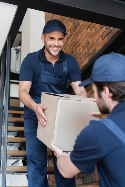 Sonriente mover llevar caja de cartón con compañero borroso mientras camina arriba - foto de stock