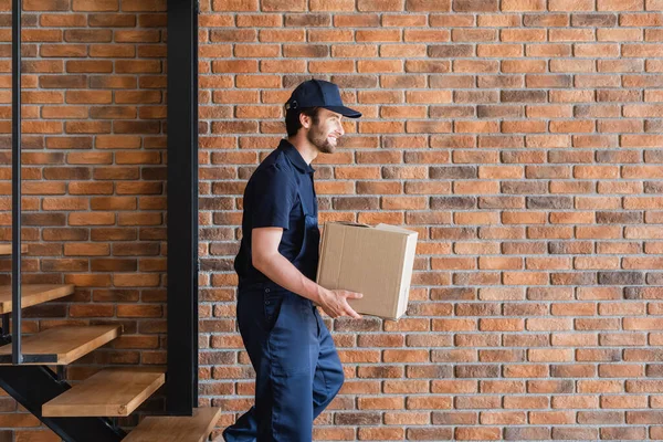 Vista lateral del movimiento sonriente que lleva la caja de cartón mientras camina abajo - foto de stock