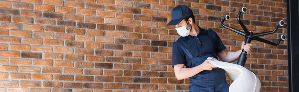 Operaio in berretto e maschera medica che tiene sedia da ufficio vicino al muro di mattoni, banner — Foto stock