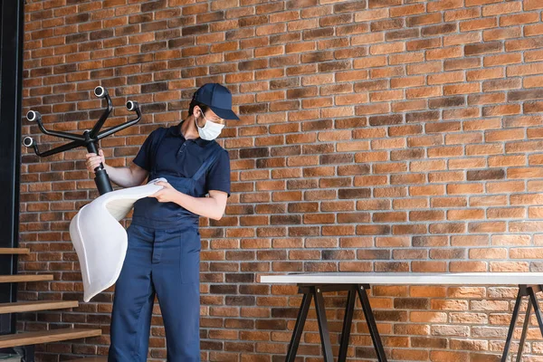 Работник в защитной маске держит офисное кресло возле кирпичной стены и стола — стоковое фото