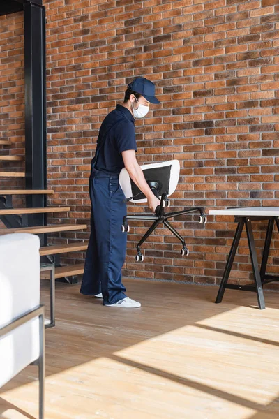 Beweger mit medizinischer Maske hält Bürostuhl in Wohnung nahe Treppe — Stockfoto