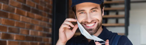 Obrero alegre sonriendo a la cámara mientras se quita la máscara médica, pancarta - foto de stock