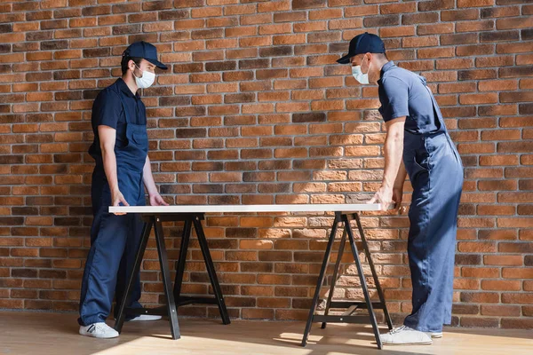 Arbeiter in Overalls und medizinischen Masken halten Sperrholz in der Nähe der Ziegelmauer — Stockfoto