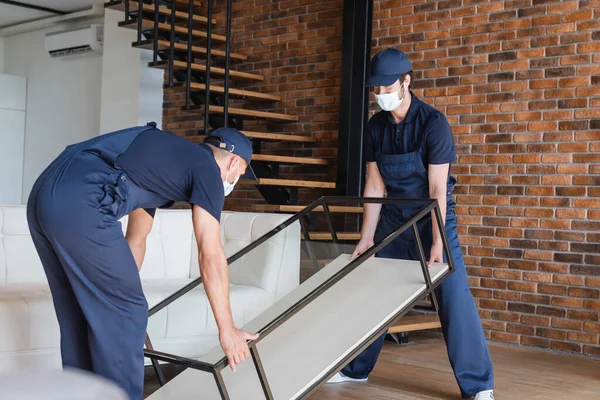 Déménageurs en uniforme et masques médicaux table de levage de verre près des escaliers — Photo de stock