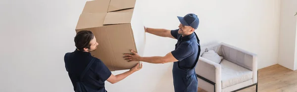 Vista de ángulo alto de los motores sonrientes que sostienen la caja de cartón en la sala de estar, pancarta - foto de stock