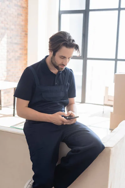 Traslocatore in tuta da lavoro messaggistica sul cellulare sul divano in appartamento — Foto stock