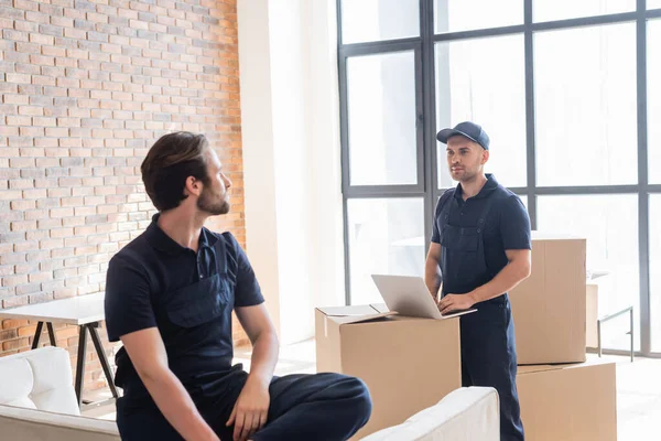 Mover tippt auf Laptop neben Kartons und Kollege auf Couch — Stockfoto