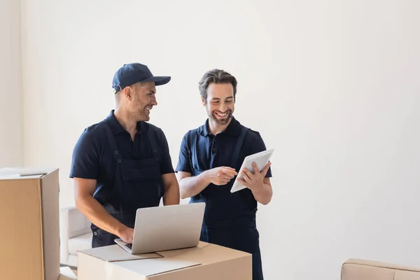 Cheerful workman pointing at digital tablet while his colleague typing on laptop near carton boxes — Stock Photo