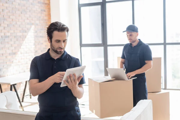 Workman using digital tablet near carton boxes and blurred colleague typing on laptop — Stock Photo