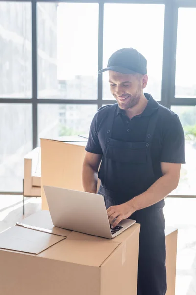 Щасливий працівник, який друкує на ноутбуці на картонній коробці біля вікон — стокове фото