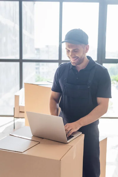 Trabajador feliz escribiendo en el ordenador portátil en caja de cartón cerca de ventanas - foto de stock