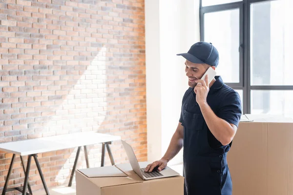 Homme positif en uniforme parlant sur smartphone tout en tapant sur ordinateur portable sur boîte en carton — Photo de stock