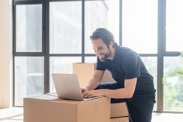 Heureux déménageur dactylographier sur ordinateur portable près de boîtes en carton — Photo de stock