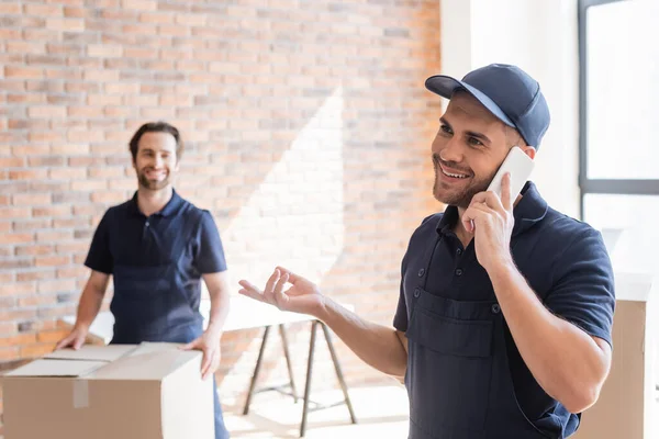 Smiling mover talking on mobile phone near blurred colleague and carton boxes — Stock Photo