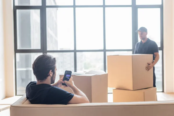 KYIV, UKRAINE - JUNE 24, 2021: mover sitting on couch and messaging on smartphone near blurred colleague and carton packages — Stock Photo