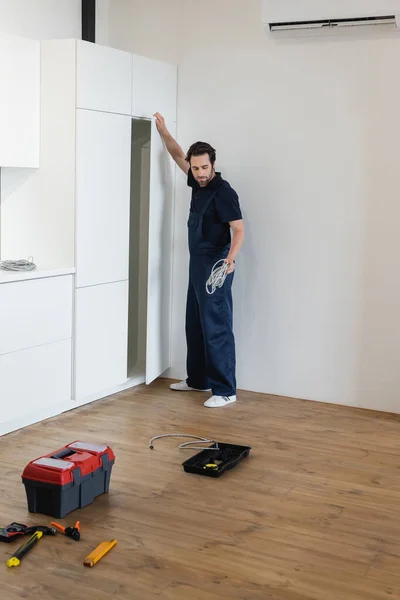 Contremaître en uniforme tenant le câble tout en se tenant près des meubles de cuisine blancs — Photo de stock