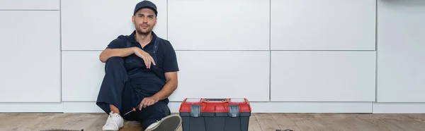 Réparateur avec tournevis assis sur le sol dans la cuisine près de la boîte à outils, bannière — Photo de stock