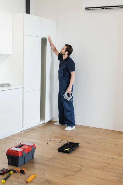 Obrero abriendo gabinete de cocina mientras sostiene el cable cerca de herramientas y caja de herramientas en el piso - foto de stock