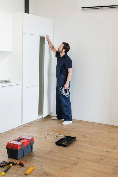 Arbeiter öffnet Küchenschrank und hält Kabel in der Nähe von Werkzeug und Werkzeugkiste auf dem Boden — Stockfoto