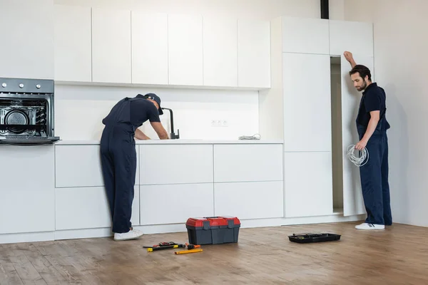 Encanador e eletricista trabalhando na cozinha moderna perto de ferramentas no chão — Fotografia de Stock