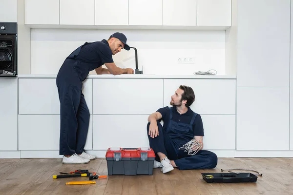 Idraulico guardando operaio seduto sul pavimento della cucina vicino cassetta degli attrezzi e strumenti — Foto stock