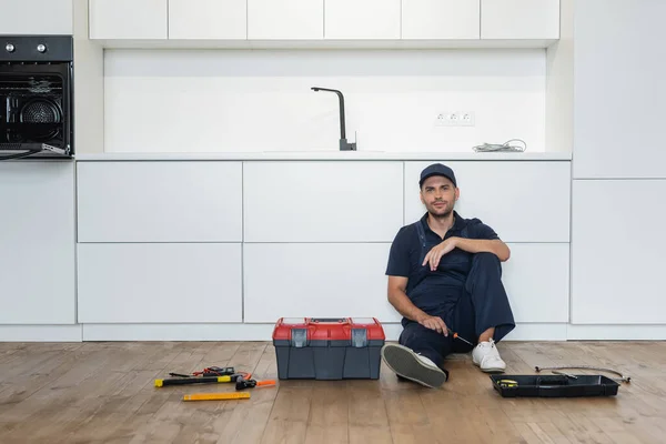 Handyman in overalls sitting on floor in kitchen near tools and toolbox — Stock Photo