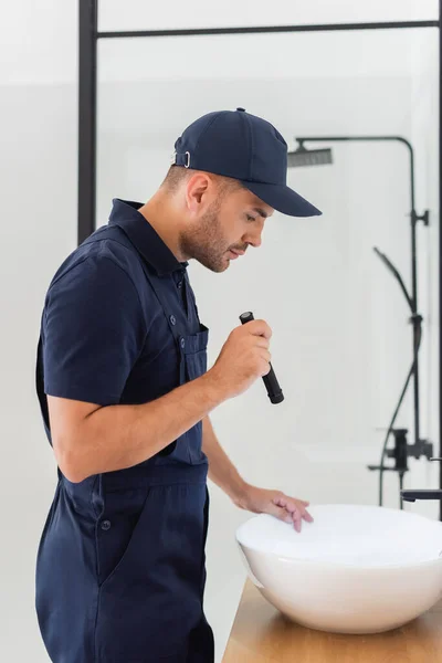 Side view of plumber with flashlight near sink in modern bathroom — Stock Photo