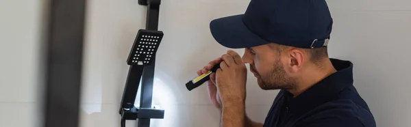 Plumber in cap holding flashlight near shower head in bathroom, banner — Stock Photo