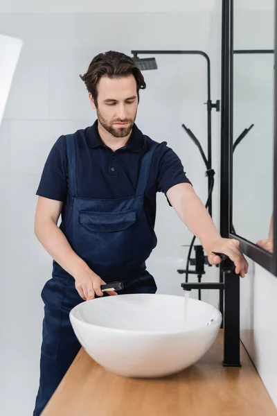 Young plumber with flashlight checking sink and faucet in bathroom — Stock Photo