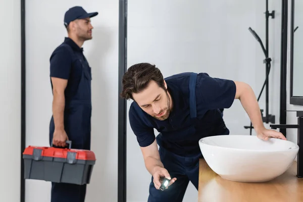 Fontanero con fregadero de control linterna en el baño cerca de colega borrosa con caja de herramientas - foto de stock