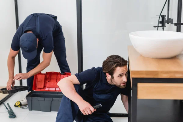 Plumber with flashlight checking sink near colleague with tools in bathroom — Stock Photo