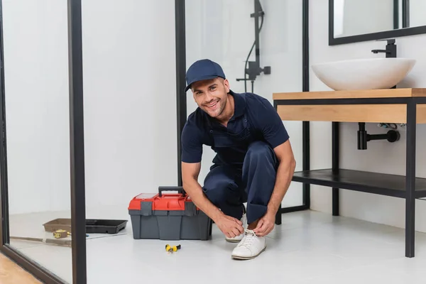 Fröhlicher Klempner bindet Schnürsenkel an Turnschuh im Badezimmer neben Werkzeugkiste auf dem Boden — Stockfoto