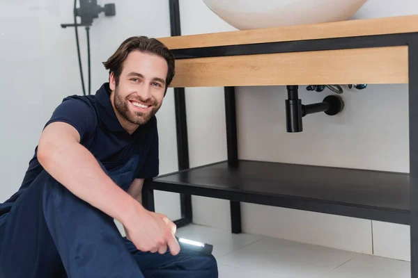 Happy plumber with flashlight looking at camera while sitting in bathroom — Stock Photo