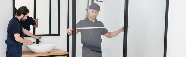 Trabalhador medindo cabine de chuveiro perto do encanador verificando pia no banheiro, banner — Fotografia de Stock
