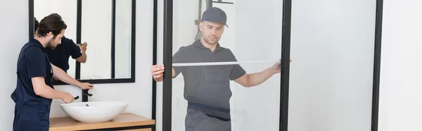 Ouvrier mesurant cabine de douche près du plombier contrôle lavabo dans la salle de bain, bannière — Photo de stock