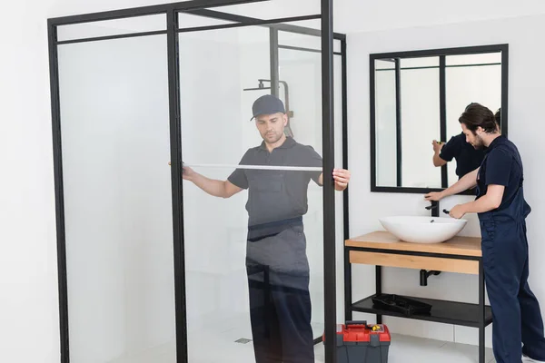 Foreman measuring shower cabin near plumber working with sink in bathroom — Stock Photo