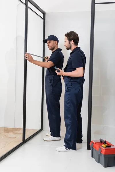 Handyman looking at colleague measuring shower cabin in bathroom — Stock Photo