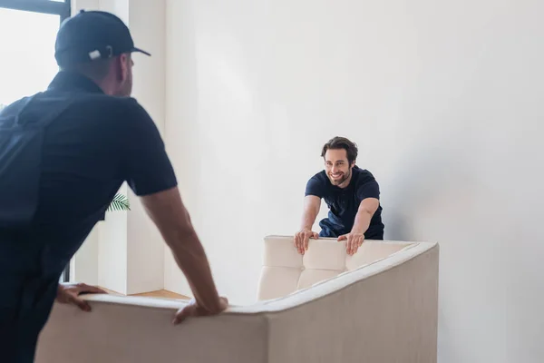 Sonriente trabajador moviendo sofá blanco con compañero borroso - foto de stock