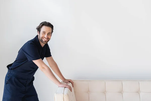 Heureux déménageur souriant à la caméra tout en se tenant près du canapé blanc — Photo de stock